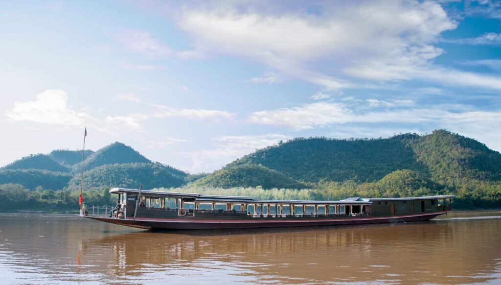 Long wooden passenger boat sailing on a calm brown river with green hills and a blue sky in the background.