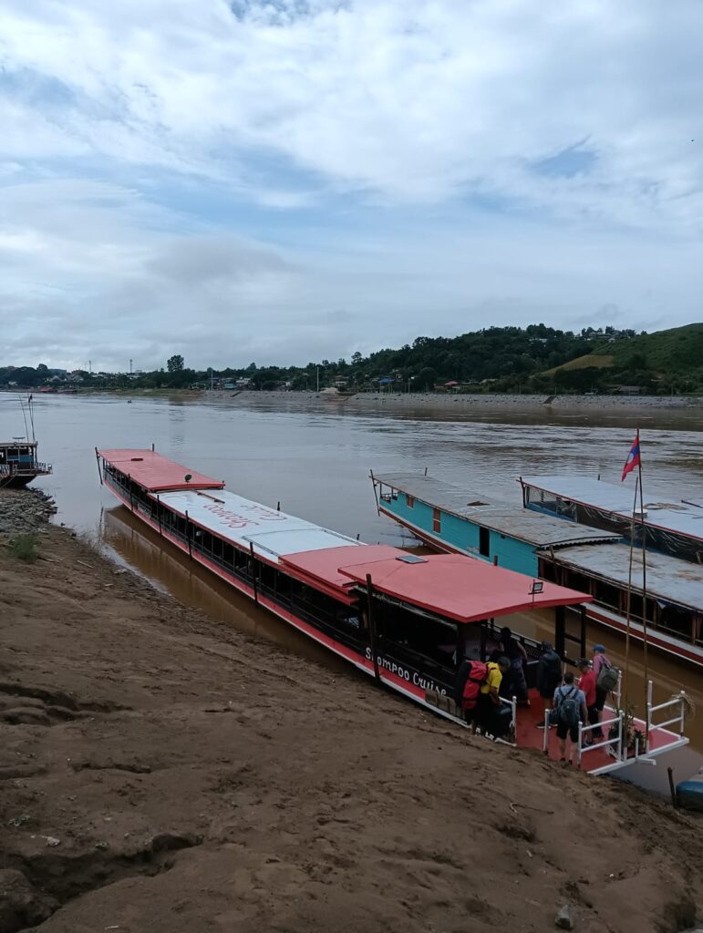 Passenger boats docked at a muddy riverbank with people boarding a red-roofed boat; a calm river and hillside village visible in the distance.