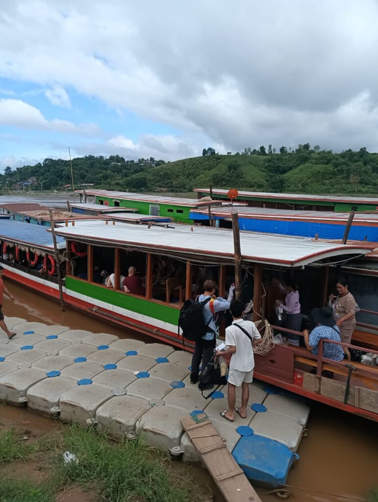 People board a long, colorful river boat at a floating dock by muddy water, with green hills in the background and a cloudy sky.