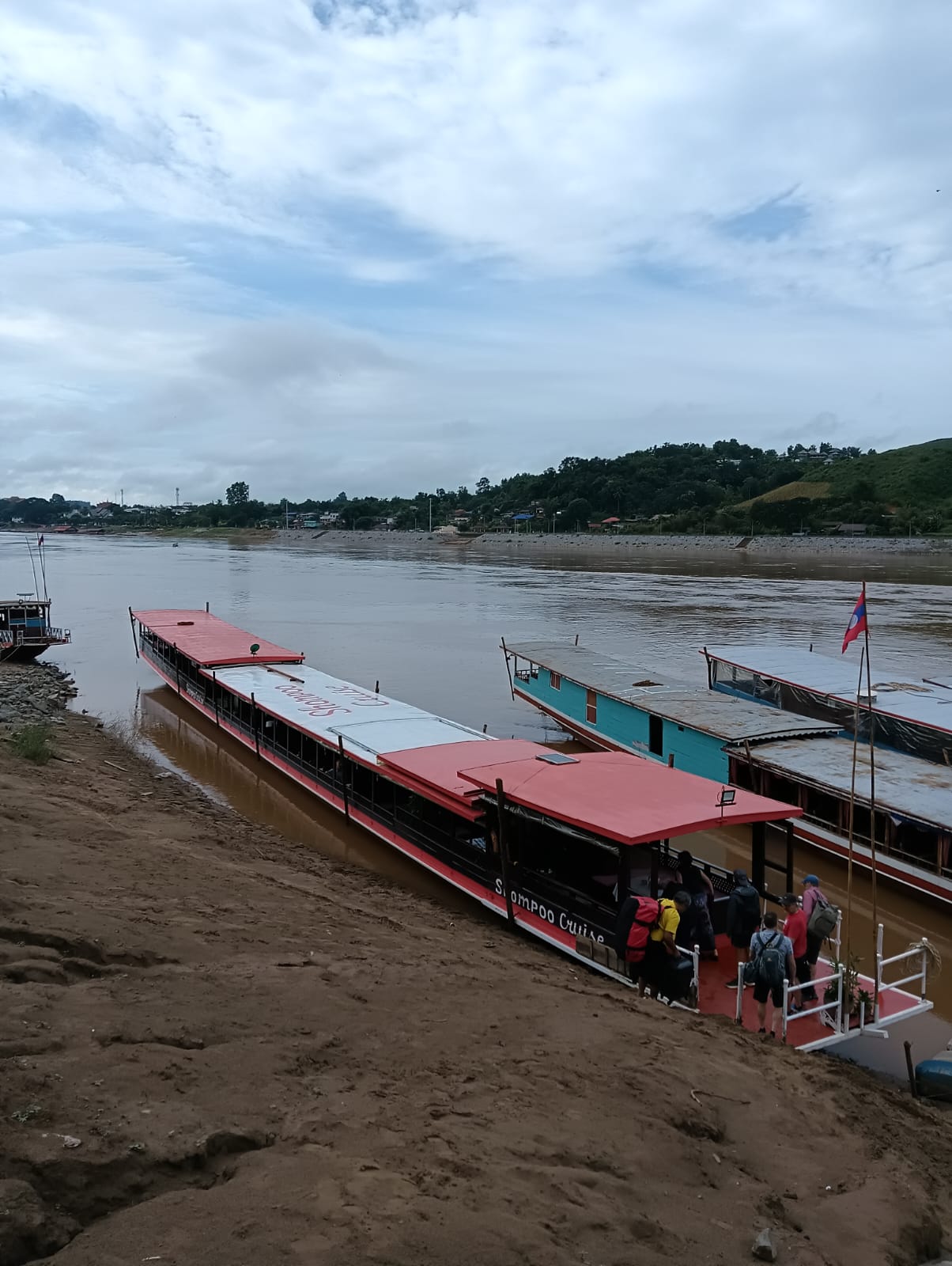 Passenger boats docked at a muddy riverbank with people boarding a red-roofed boat; a calm river and hillside village visible in the distance.